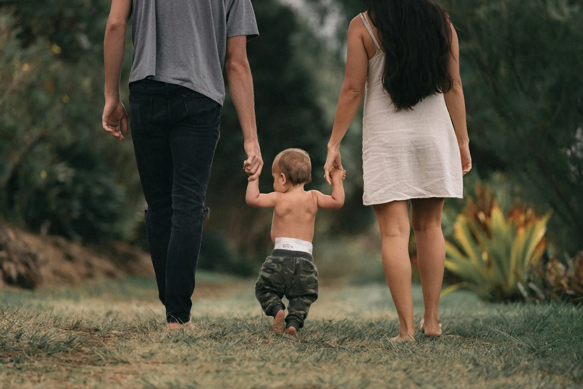 Parents holding a toddler's hands as he walks between them on the grass